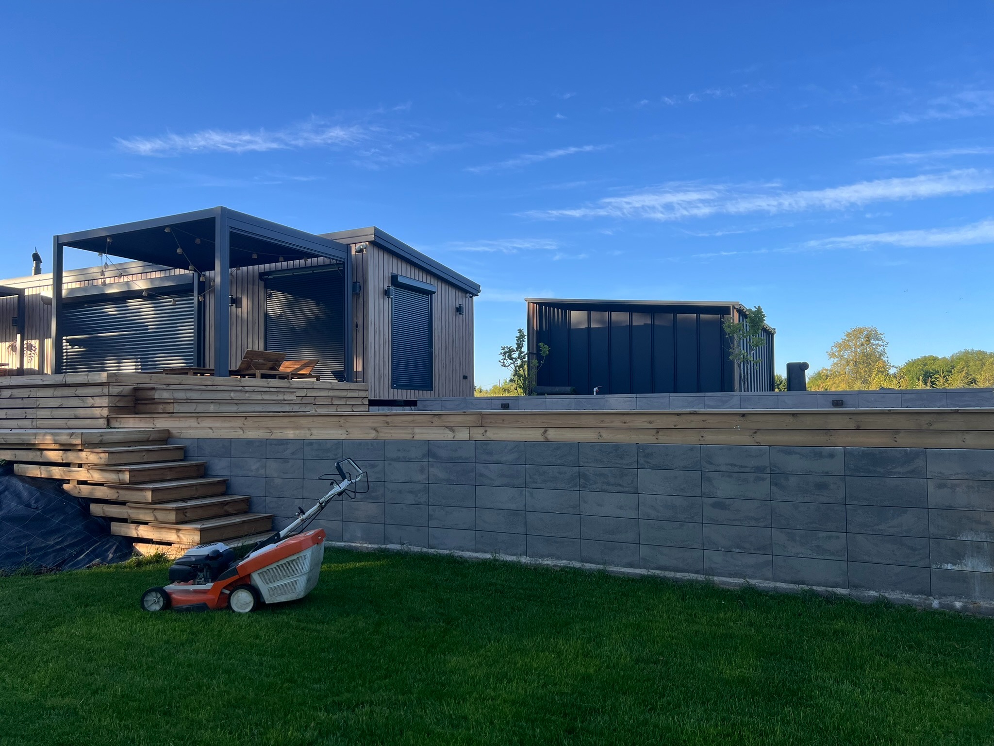Nowoczesny ogród z pergolą, drewnianymi schodami i równo skoszoną trawą. W tle widoczny budynek. Na pierwszym planie kosiarka do trawy.