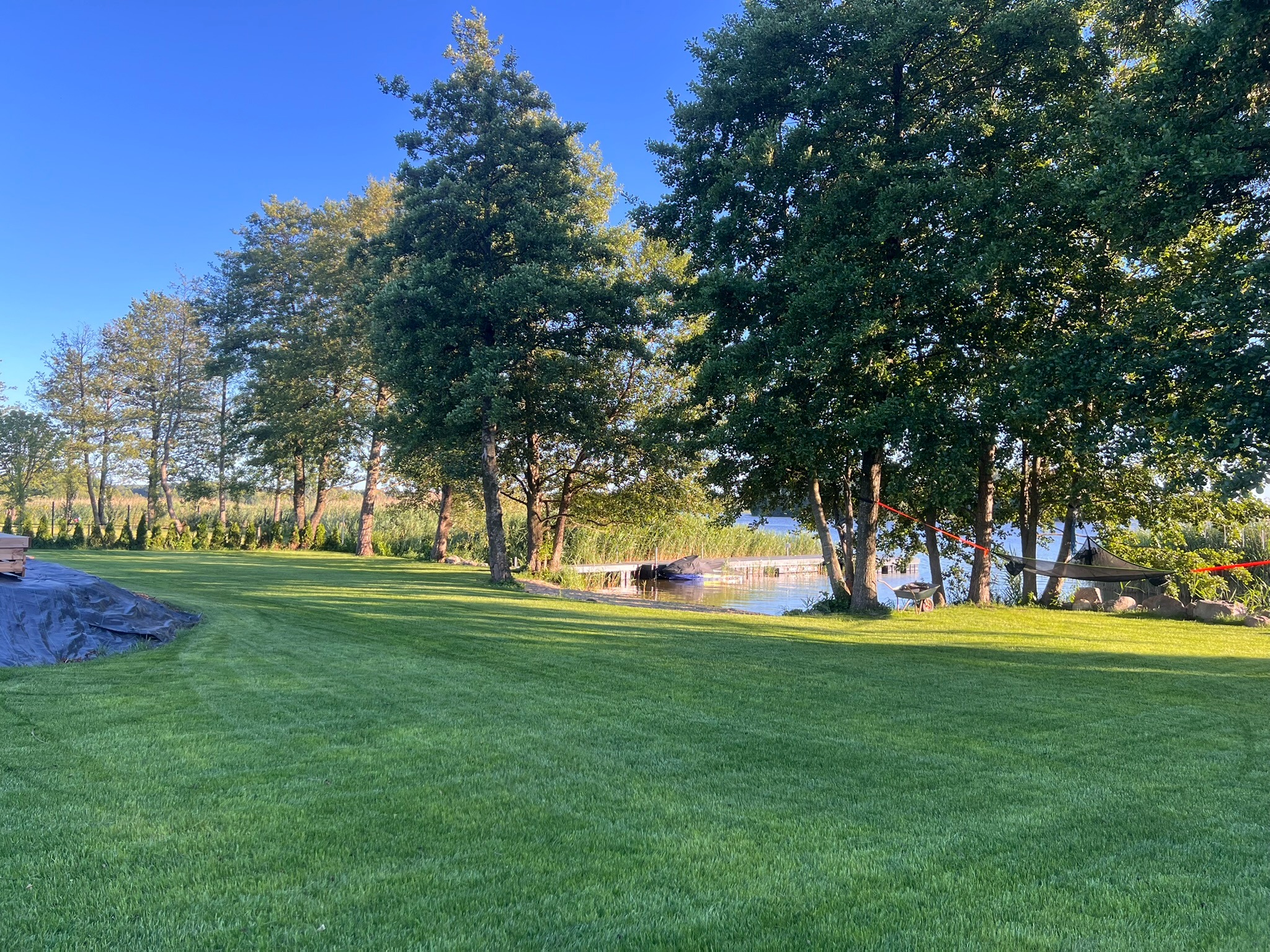 Rozległy, zielony trawnik z widokiem na jezioro i pomost. W tle drzewa, a w lewym rogu materiał budowlany przykryty folią. Słoneczny dzień.