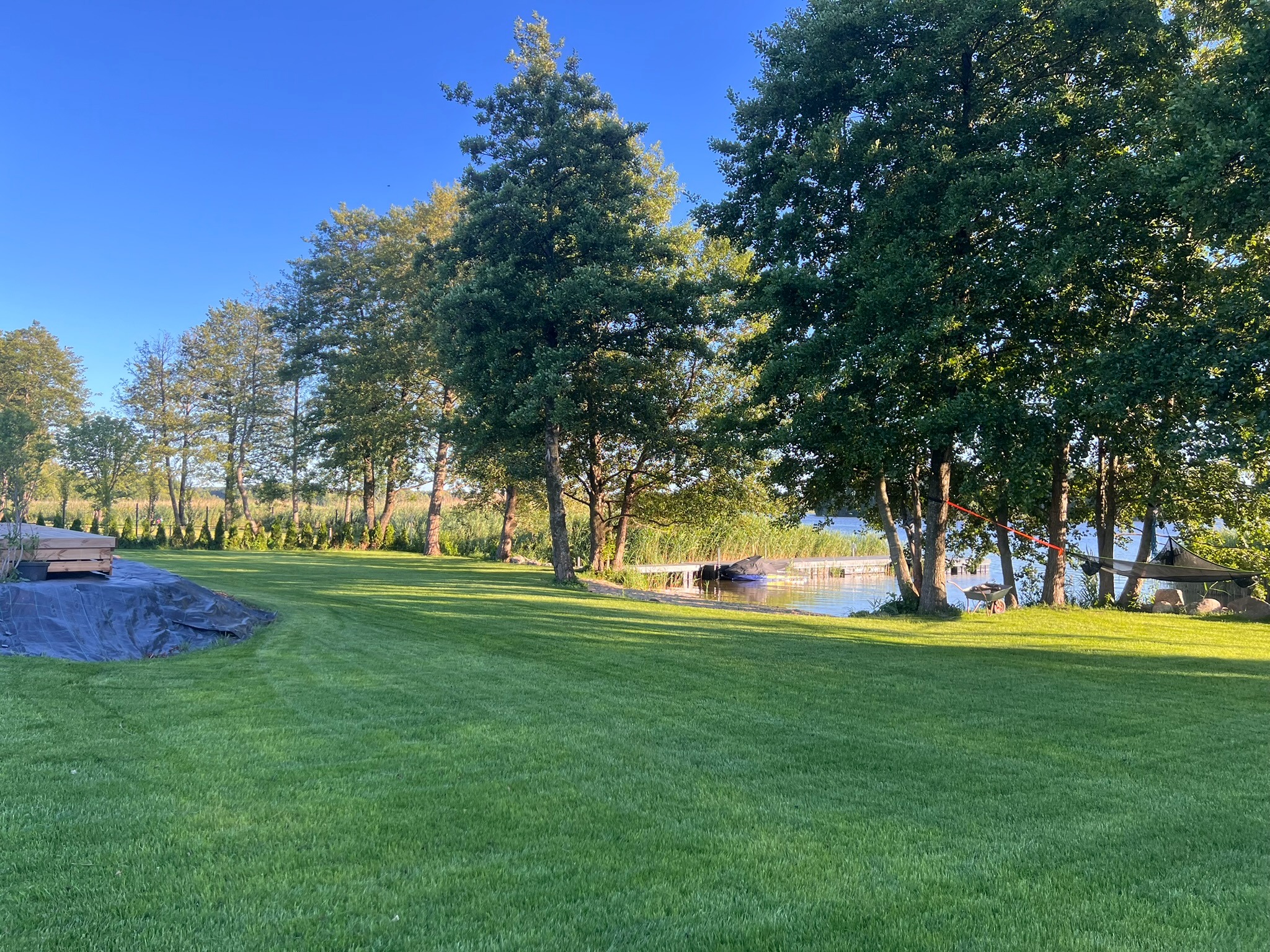 Rozległy trawnik nad jeziorem w słoneczny dzień. Widoczny pomost, drzewa i przygotowane podłoże pod nasadzenia, co sugeruje prace związane z zagospodarowaniem terenu.