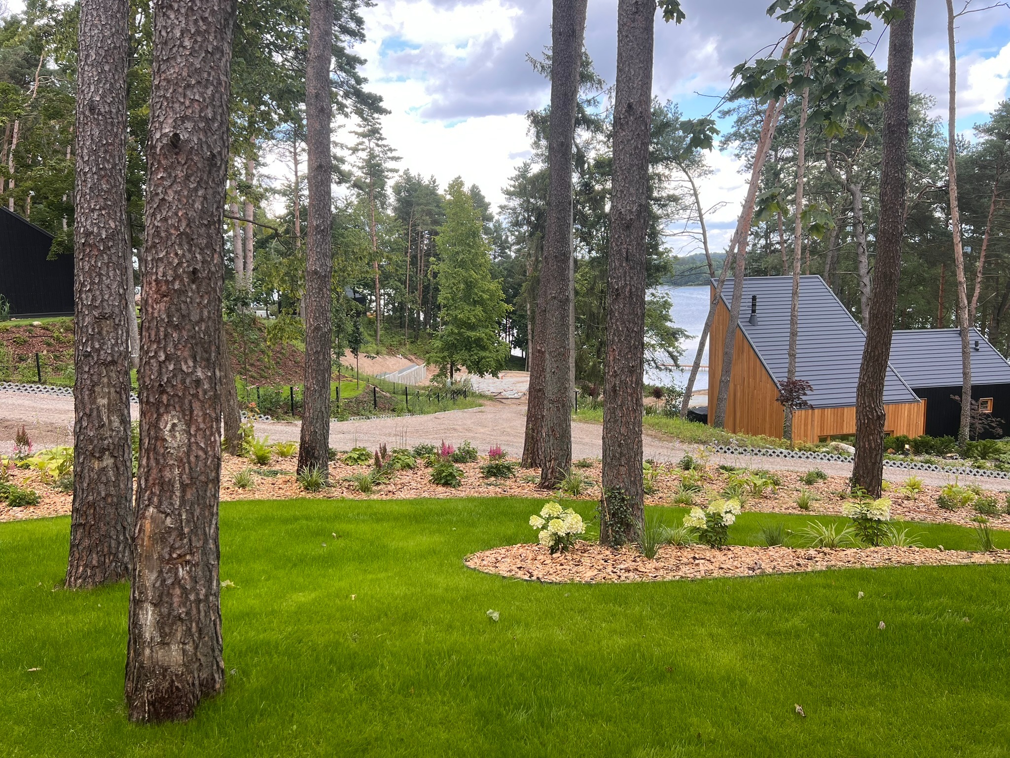 Malowniczy widok na świeżo założony ogród z równo przystrzyżonym trawnikiem, rabatami kwiatowymi i korą wokół drzew, z domem w tle nad jeziorem.
