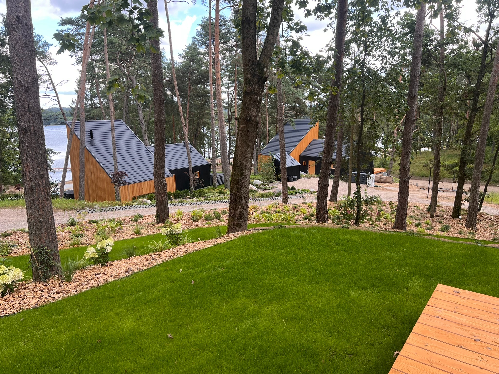 Nowoczesne domki letniskowe z ciemnymi dachami i drewnianą elewacją, otoczone zielonym trawnikiem i drzewami, widok z perspektywy trawnika.