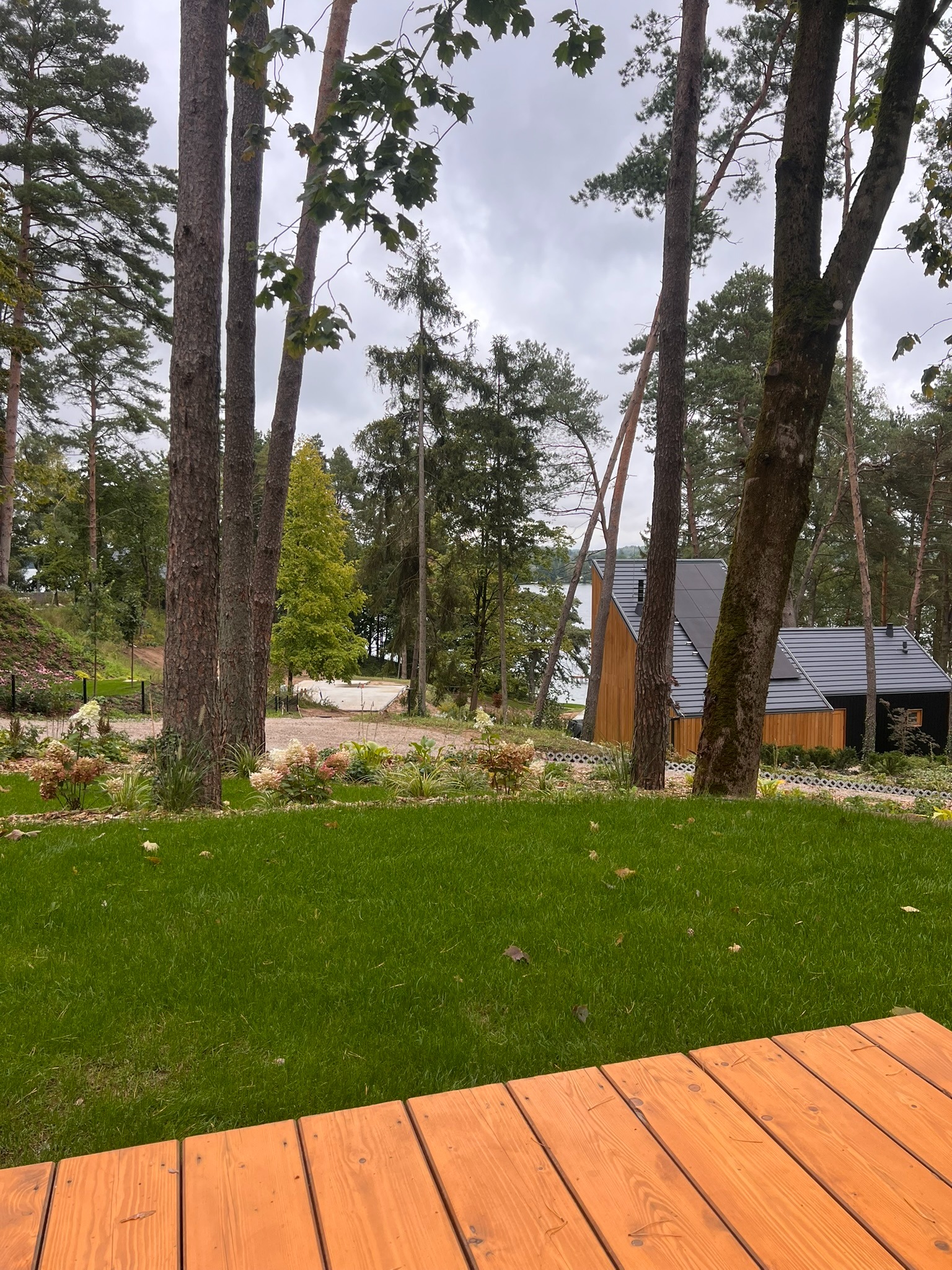 Widok na nowoczesny dom z panelami słonecznymi, otoczony zielenią i drzewami, z perspektywy drewnianego tarasu. Uwagę przykuwa zadbany trawnik i kompozycja roślin.