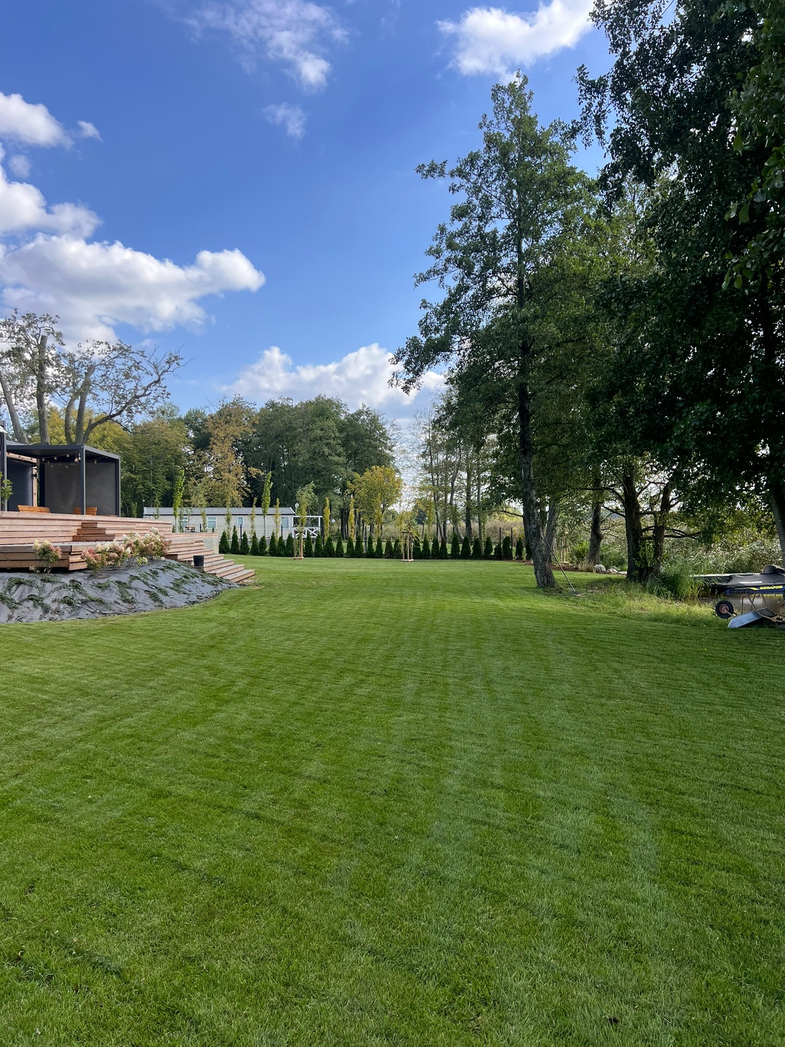 Perfekcyjnie utrzymany trawnik z równo przystrzyżoną trawą, otoczony drzewami i elementami architektury ogrodowej w słoneczny dzień. Widoczny fragment tarasu.