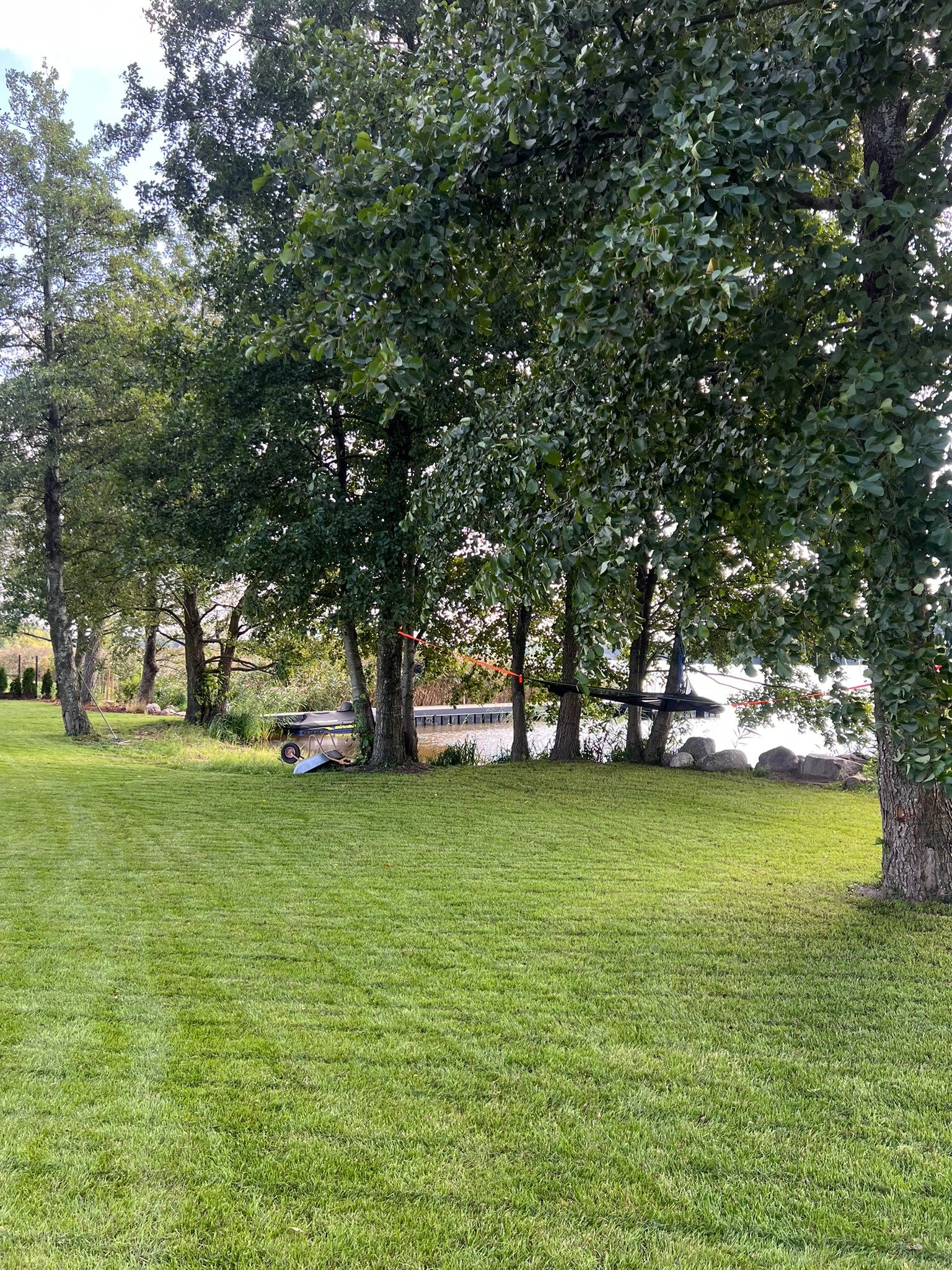 Zadbany trawnik z widocznymi pasami po koszeniu, drzewa liściaste w tle, w oddali pomost nad wodą. Wśród drzew widoczny wózek ogrodowy.