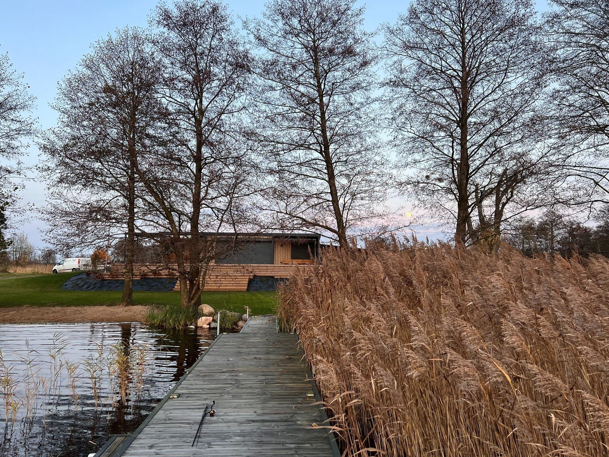 Drewniany pomost nad jeziorem z trzcinami, prowadzący do nowoczesnego domu z elementami drewnianymi. W tle drzewa bez liści i błękitne niebo o zmierzchu.