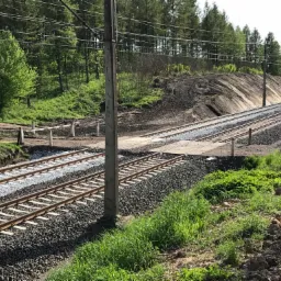 Nasyp kolejowy z dwoma torami po renowacji, świeżo wysypany tłuczeń i podsypka, widoczne słupy trakcyjne i fragment barierek zabezpieczających.