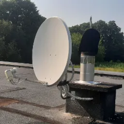 Biała antena satelitarna zamontowana na kominie wentylacyjnym na dachu budynku, z widokiem na drzewa i budowę w tle.