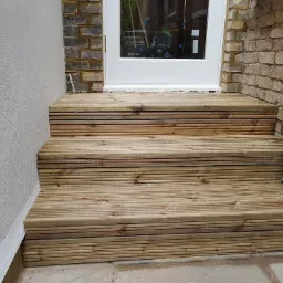 Trzy stopnie schodów z desek tarasowych prowadzące do białych drzwi na tle ściany z jasnej cegły i szarego tynku.