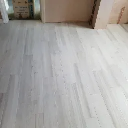 Ułożona podłoga z jasnych paneli podłogowych imitujących drewno, widoczne kartony w przejściu do innego pomieszczenia, ściany w trakcie wykańczania.