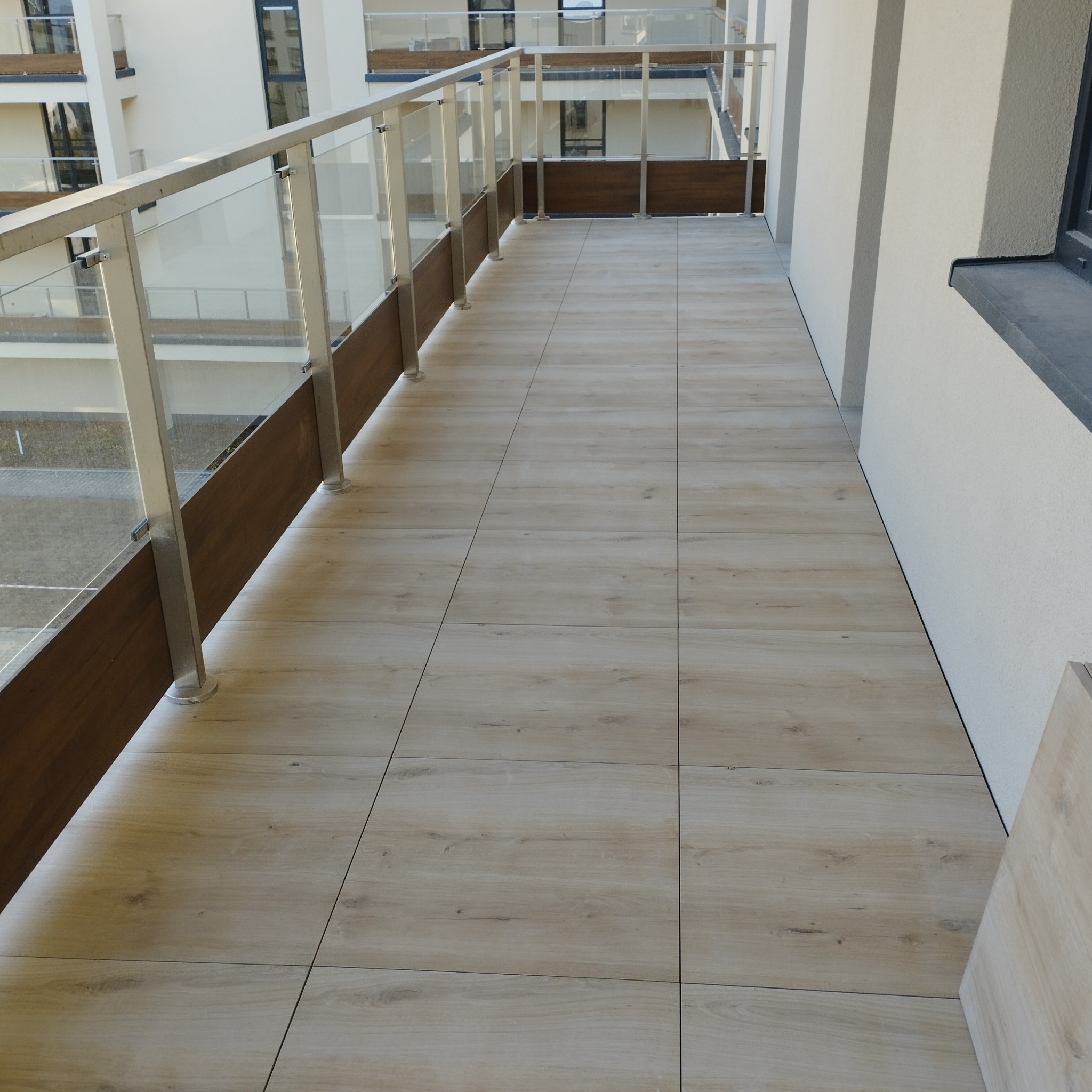 Wykończony balkon w Kielcach z podłogą wyłożoną jasnymi płytkami imitującymi drewno, szklane balustrady z drewnianymi elementami wykończeniowymi.