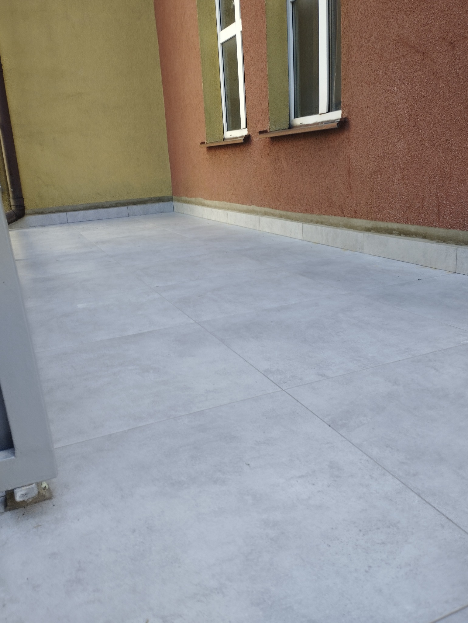 Wyłożony jasnoszarymi płytkami taras przylegający do ściany budynku z dwoma oknami, widoczne fugi i listwa przypodłogowa.
