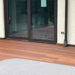 Nowoczesny taras z egzotycznego drewna, przylegający do budynku z dużymi przesuwnymi drzwiami, widoczny fragment szarego mebla ogrodowego.