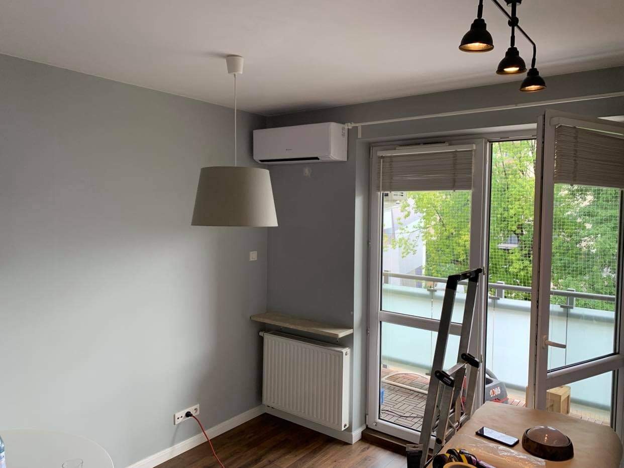 Klimatyzator ścienny zamontowany w szarym pokoju z widokiem na balkon, drabina oparta o okno balkonowe, widoczne narzędzia na stole.