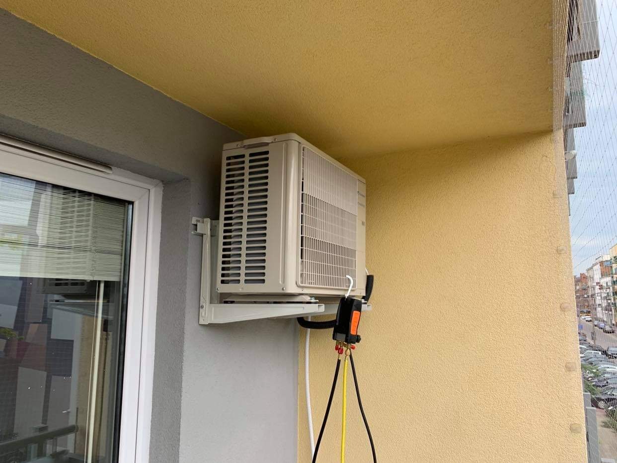 Zewnętrzna jednostka klimatyzatora zamontowana na balkonie bloku mieszkalnego, podłączona do narzędzi serwisowych z przewodami, widok z boku, siatka zabezpieczająca balkon w tle.