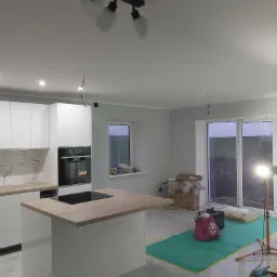 Wnętrze kuchni w trakcie remontu: białe szafki z marmurkowym backsplash, wyspa z drewnianym blatem i płyta indukcyjna, surowe przewody oświetleniowe zwisające z sufitu, kartony i lampa budowlana...