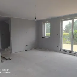 Szare, surowe wnętrze w trakcie wykańczania, widoczne gładkie ściany, wylewka podłogowa, okno z widokiem na zieleń i wisząca żarówka na kablu.