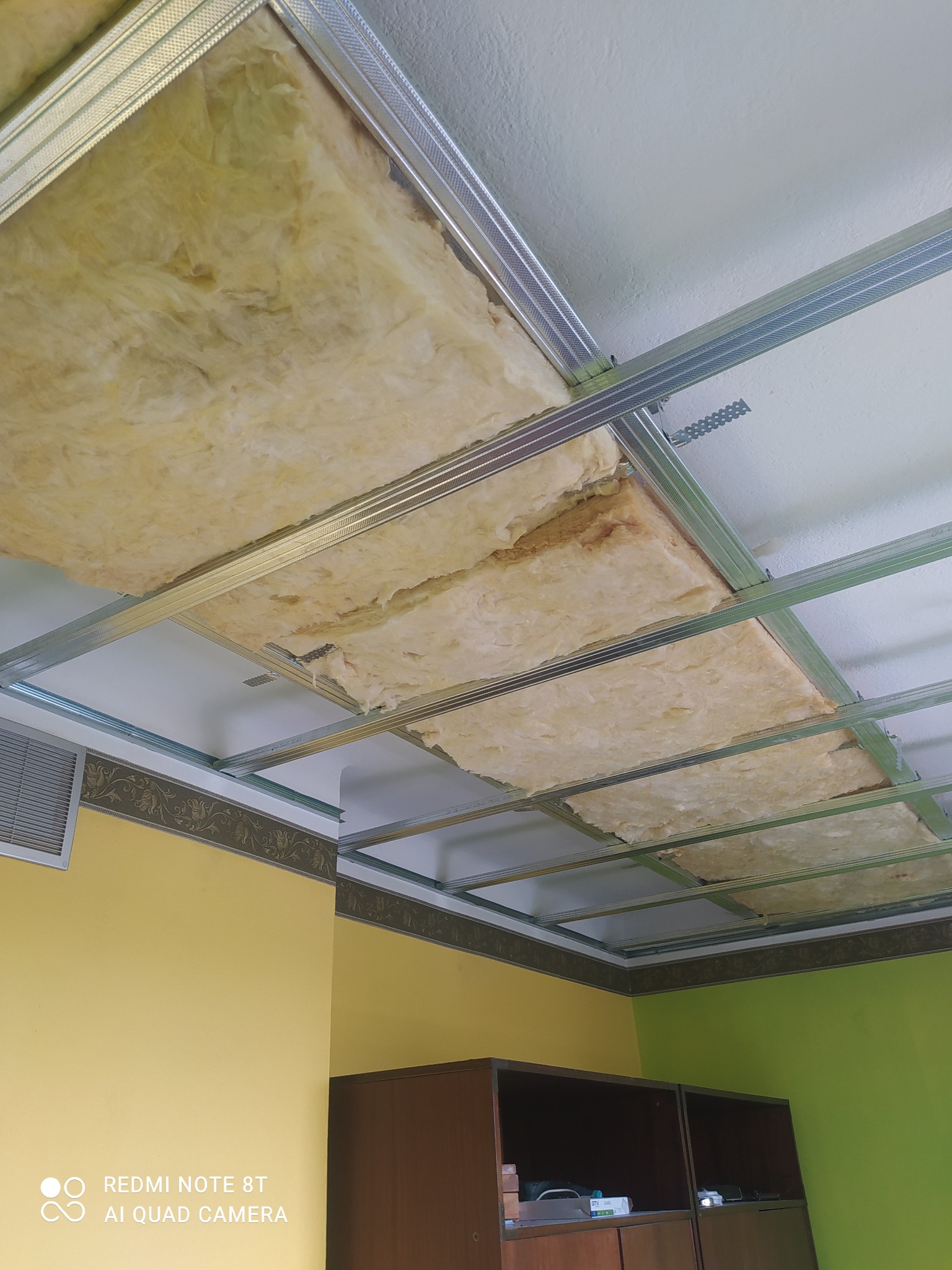 Montaż sufitu podwieszanego z widoczną izolacją z wełny mineralnej w metalowej konstrukcji, żółto-zielone ściany w tle.