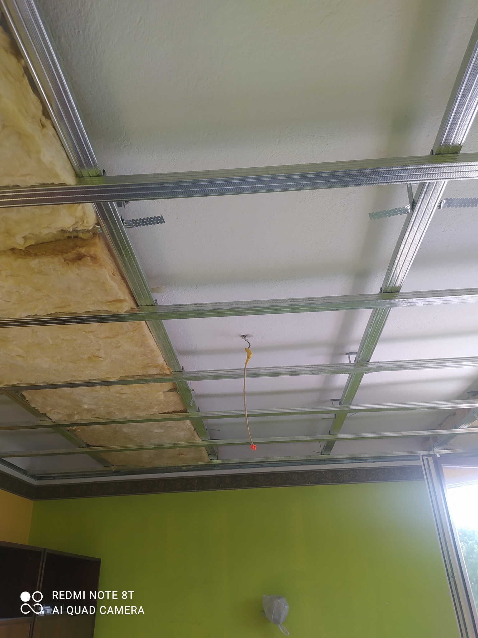 Konstrukcja metalowa sufitu podwieszanego z izolacją z wełny mineralnej, widok z dołu na tle zielonej ściany i fragmentu okna.