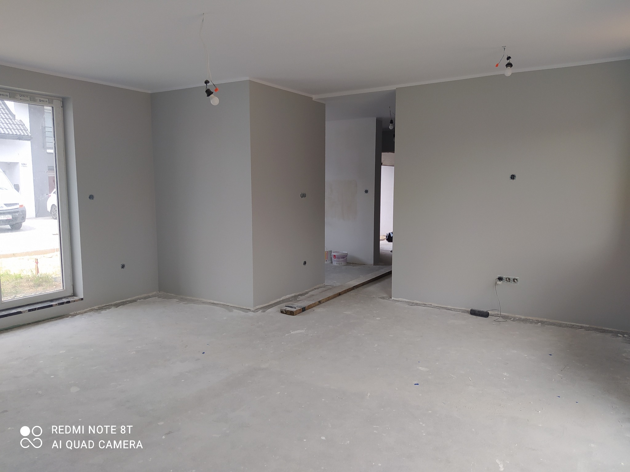 Surowe wnętrze w odcieniach szarości z widocznymi puszkami elektrycznymi, niedokończonymi wylewkami i prowizorycznym oświetleniem. W tle widoczne wiadro farby i fragment domu za oknem.