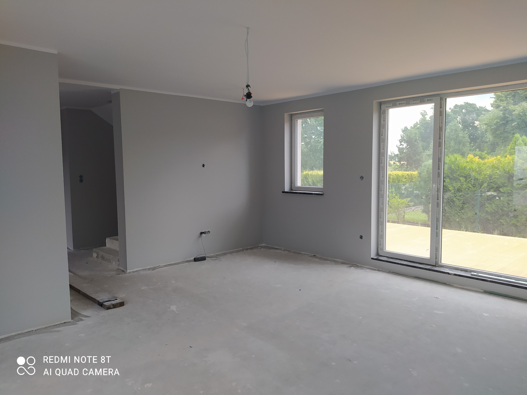 Szare, surowe wnętrze w trakcie wykańczania, widoczne gładkie ściany, wylewka podłogowa, okno z widokiem na zieleń i wisząca żarówka na kablu.