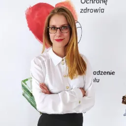 Kobieta w okularach i białej koszuli z rękami skrzyżowanymi na tle ściany z rysunkami symbolizującymi ochronę zdrowia i przyszłość.