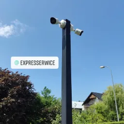 Dwie kamery monitoringu na czarnym słupie, skierowane w różnych kierunkach, na tle błękitnego nieba i zieleni.