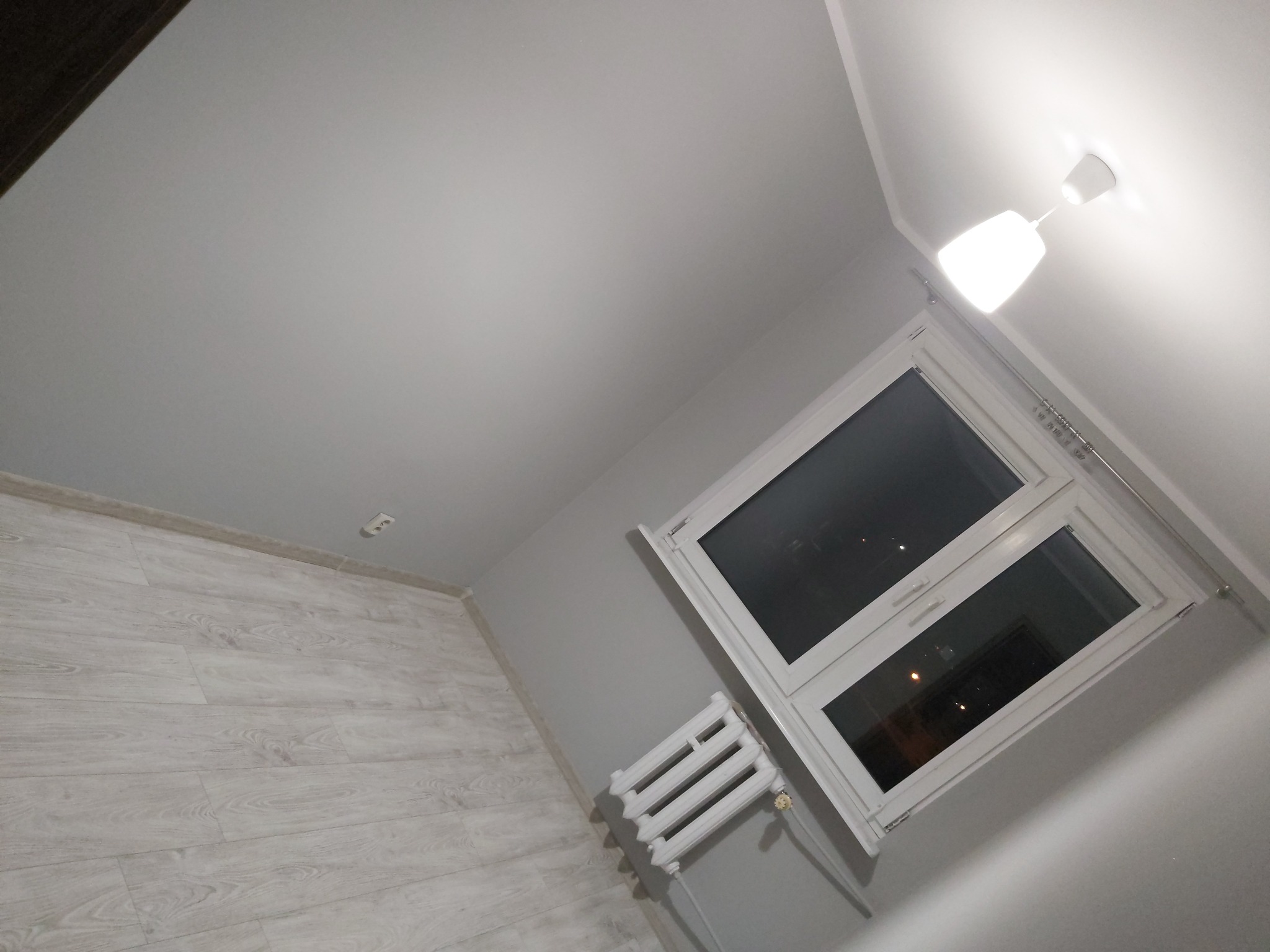Wnętrze pokoju po remoncie: jasne panele podłogowe, szare ściany, białe okno z grzejnikiem i sufit z lampą.