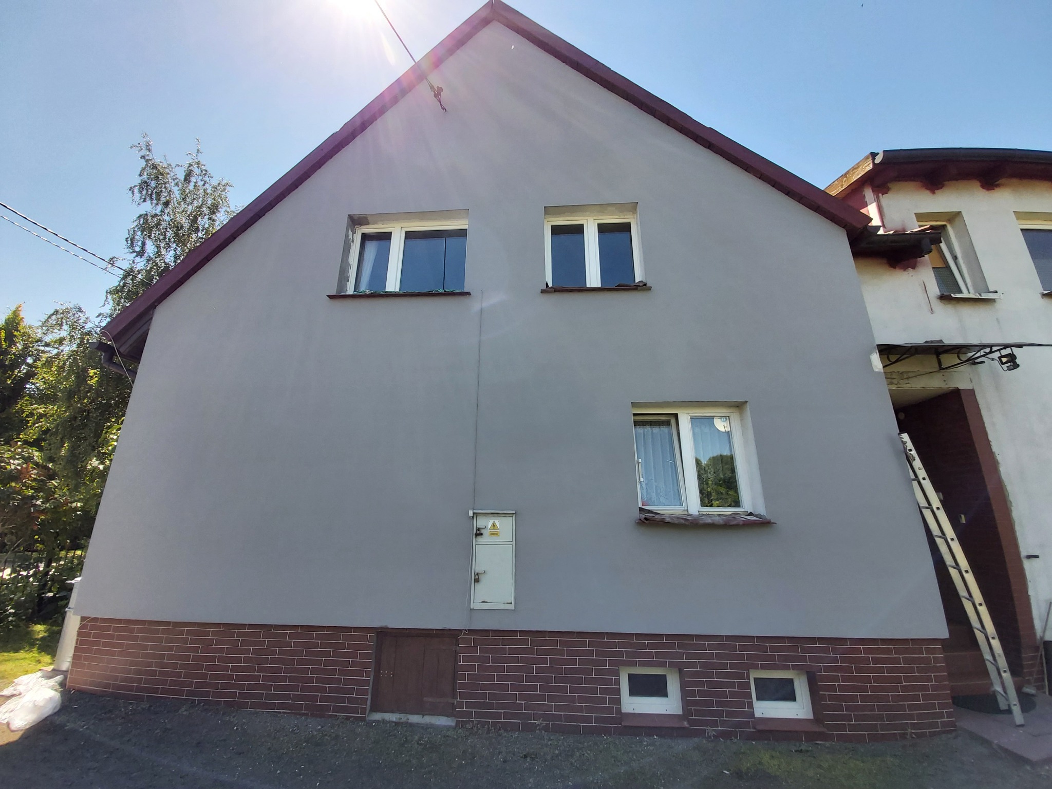 Dwupiętrowy dom z nową, szarą elewacją, widoczne dwa okna na górze i jedno na dole, ceglana podmurówka, małe drzwi techniczne i skrzynka elektryczna na elewacji, drabina oparta o budynek sąsiadujący.