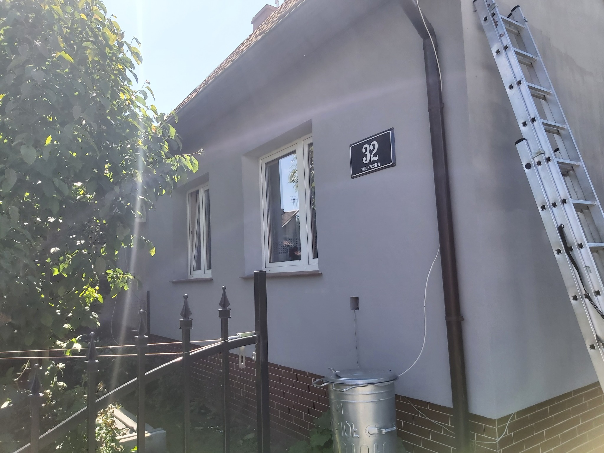 Dom jednorodzinny po tynkowaniu elewacji na szaro, widoczna drabina oparta o ścianę, adres '32 Wileńska' na tabliczce, metalowy kosz na śmieci przy ścianie i fragment ogrodzenia.