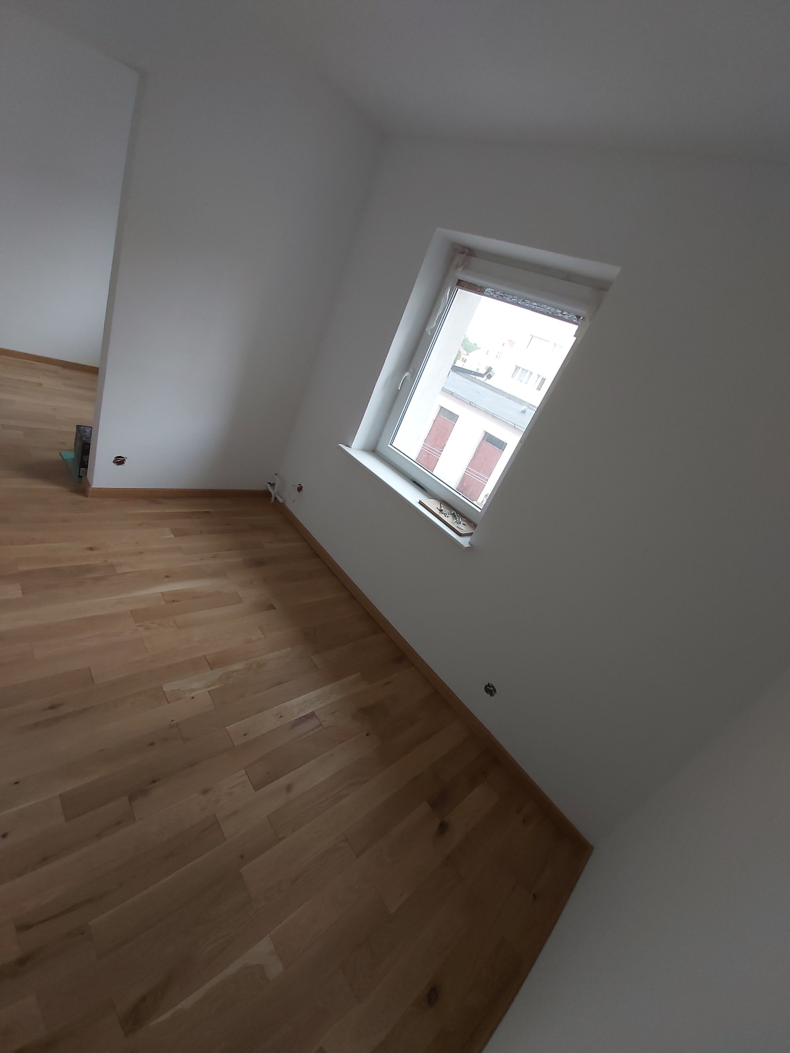 Jasny parkiet ułożony w nowym pokoju z białymi ścianami i oknem z widokiem na budynki.