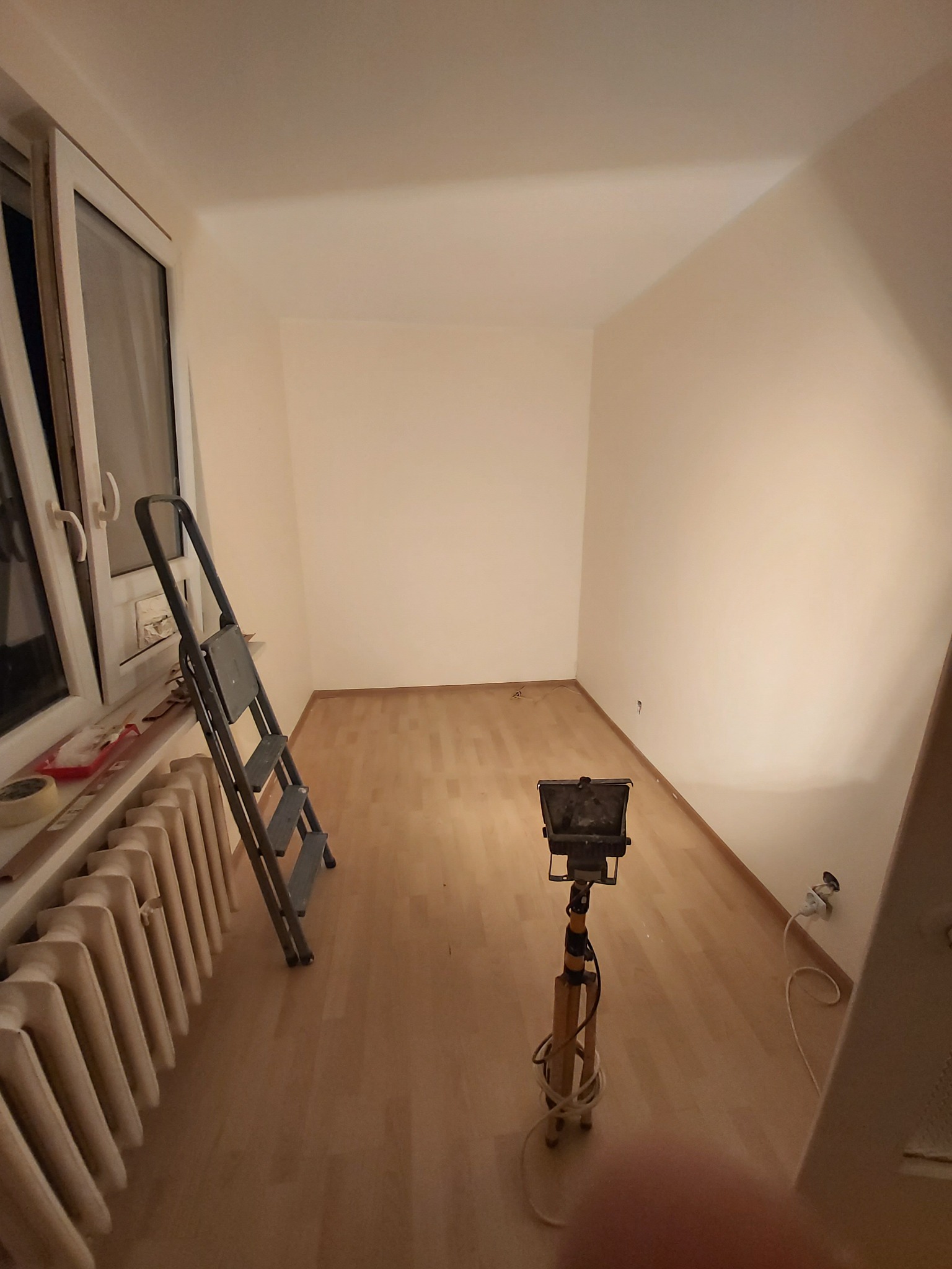 Pokój po malowaniu ścian na biało, widoczna drabina, grzejnik pod oknem, lampa budowlana na statywie i jasna drewniana podłoga.