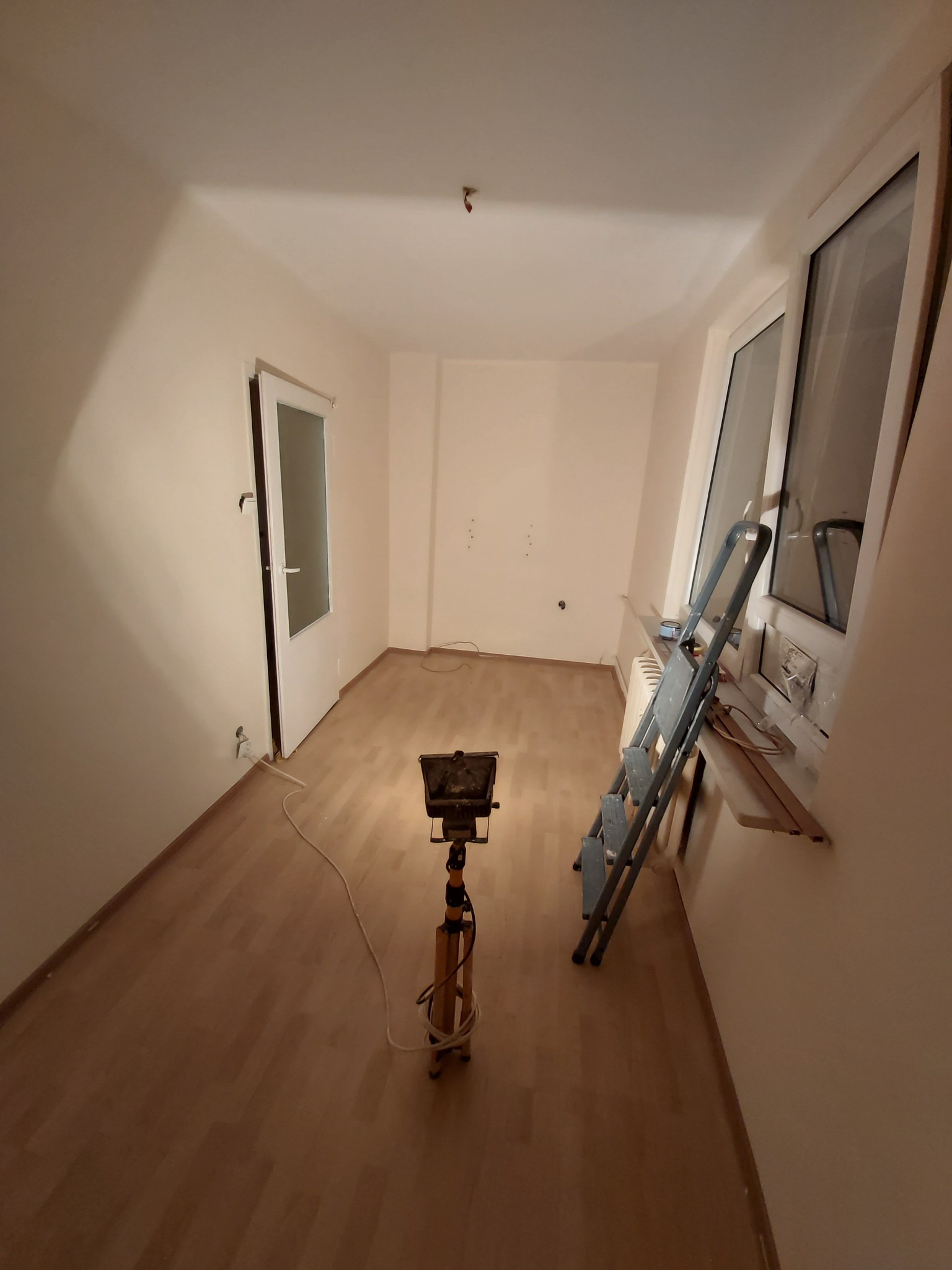 Pusta przestrzeń w trakcie remontu, z drewnianą podłogą, białymi ścianami, drabiną opartą o okno, lampą budowlaną na statywie i otwartymi drzwiami.
