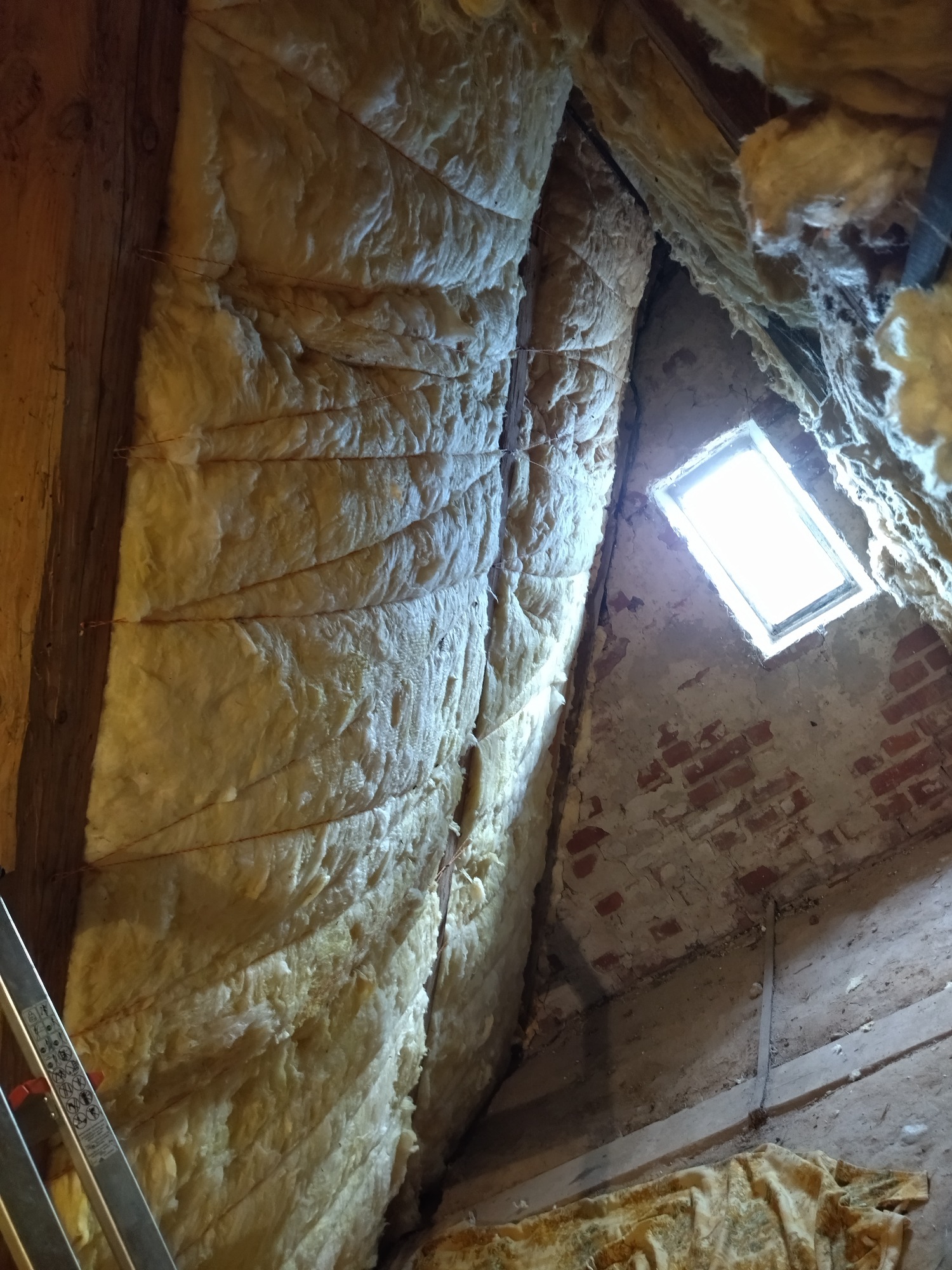 Ocieplenie poddasza wełną mineralną, widoczne drewniane elementy konstrukcyjne, ceglany fragment ściany i okno dachowe w surowym stanie.