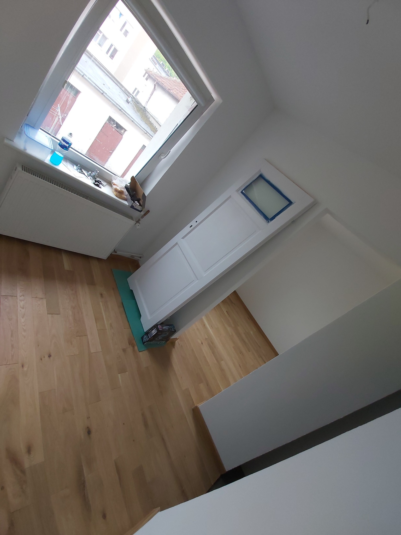 Białe drzwi z niebieską taśmą zabezpieczającą szybę, oparte o ścianę na podłodze z jasnego parkietu w trakcie remontu mieszkania, widoczny grzejnik i okno dachowe.