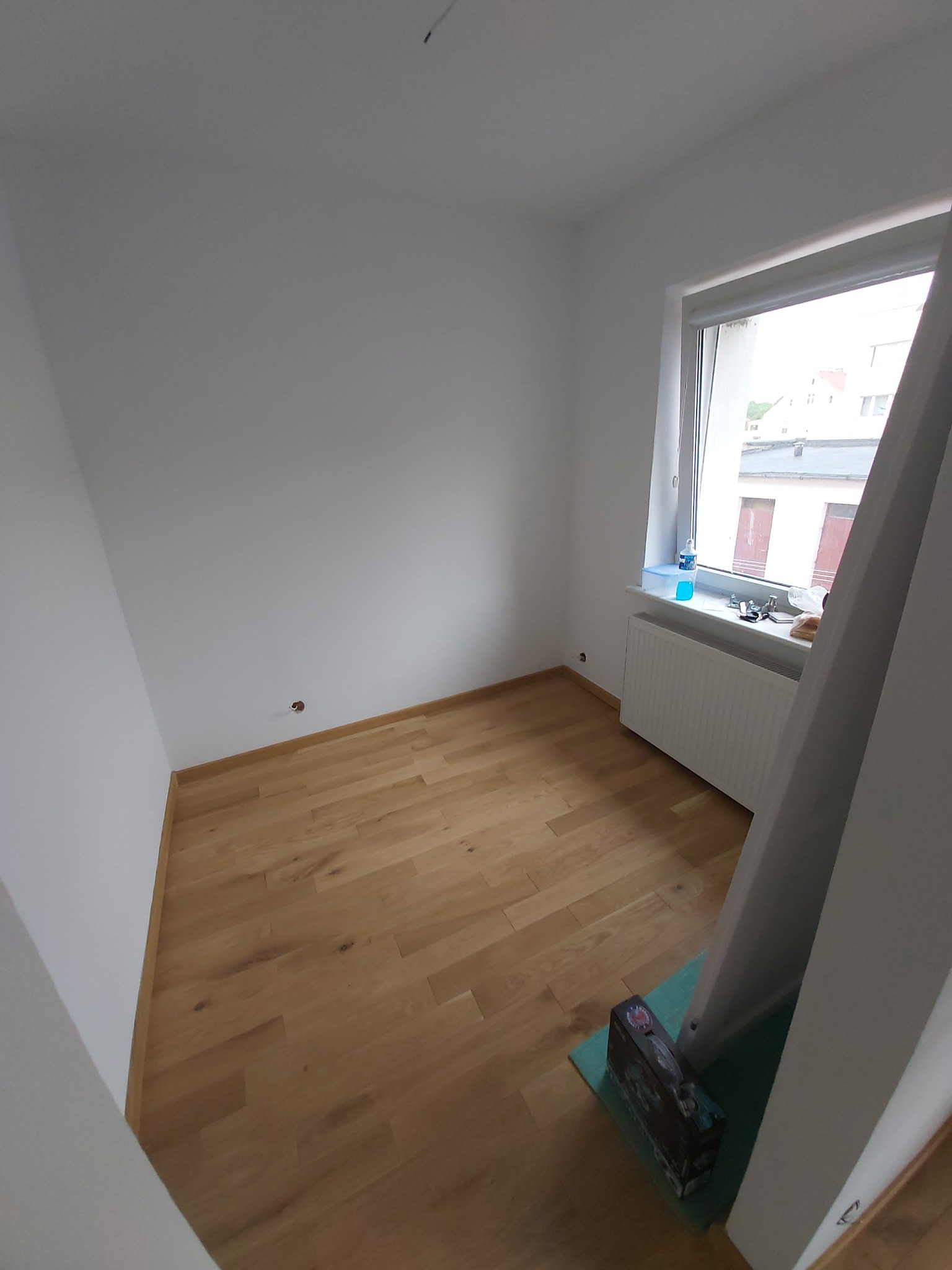 Pusty pokój po położeniu parkietu z widocznym oknem, grzejnikiem i narzędziami do montażu podłogi.
