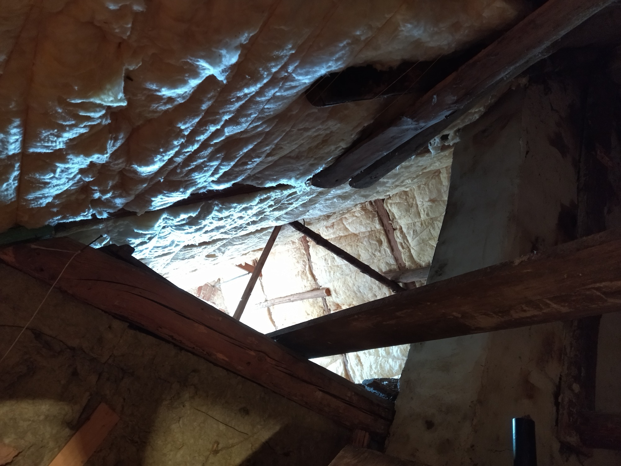 Poddasze z widoczną izolacją piankową na skośnym dachu, drewniane belki konstrukcyjne w ciemnym ujęciu, fragment muru w tle.