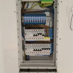 Rozdzielnica elektryczna z zainstalowanymi bezpiecznikami i modułami, widoczne kable i oznaczenia na ścianie.
