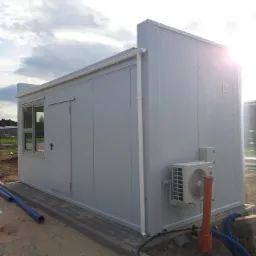 Szary kontener biurowy z klimatyzacją na zewnątrz, widoczne rury i teren w trakcie przygotowania.