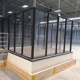 Metalkoop Bartosz Banaś - Szklana konstrukcja z czarnymi ramami, przypominająca boks lub kabinę dźwiękoszczelną, umieszczona na podwyższeniu z drewnianą obudową z żółtymi wspornikami, w przestronnym, surowym wnętrzu hali...