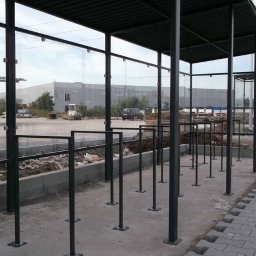 Metalkoop Bartosz Banaś - Metalowa konstrukcja z zadaszeniem i szklanymi ścianami, prawdopodobnie wiata przystankowa lub zadaszone miejsce postojowe dla rowerów, w tle budynek przemysłowy i sprzęt budowlany.