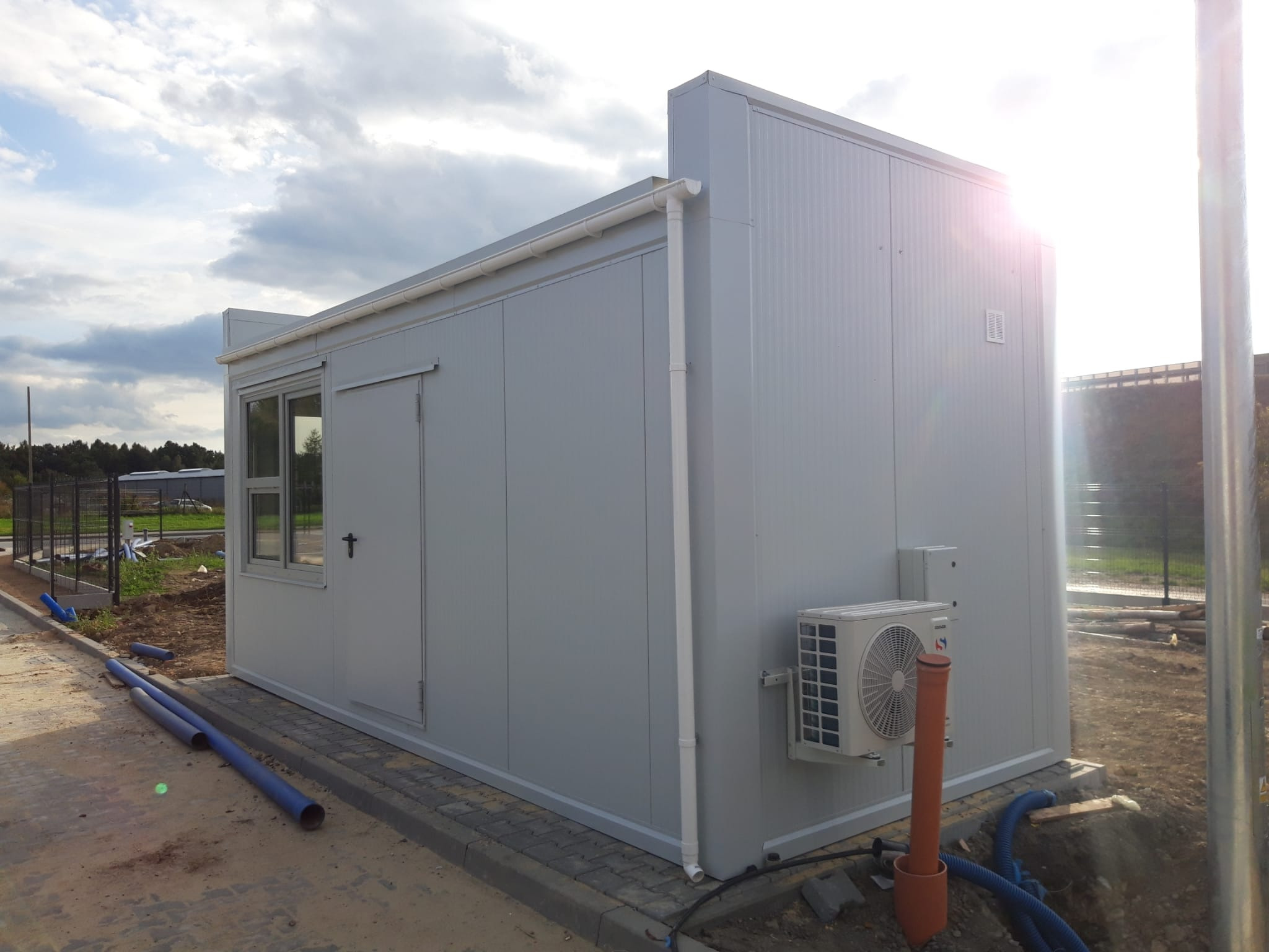 Szary kontener biurowy z klimatyzacją na zewnątrz, widoczne rury i teren w trakcie przygotowania.