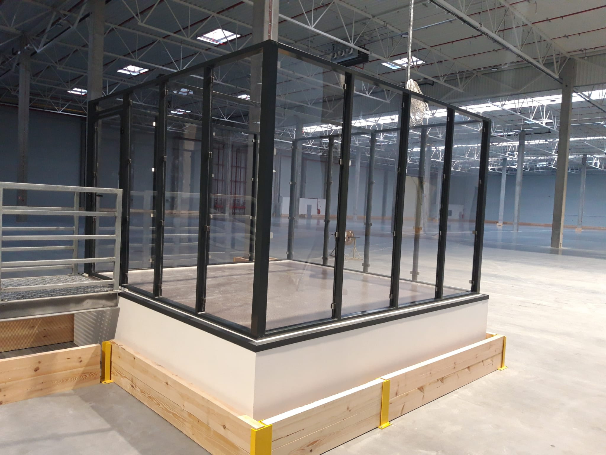 Szklana konstrukcja z czarnymi ramami, przypominająca boks lub kabinę dźwiękoszczelną, umieszczona na podwyższeniu z drewnianą obudową z żółtymi wspornikami, w przestronnym, surowym wnętrzu hali...