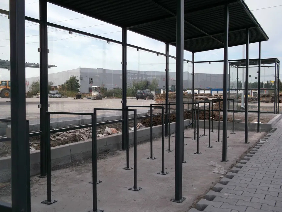 Metalowa konstrukcja z zadaszeniem i szklanymi ścianami, prawdopodobnie wiata przystankowa lub zadaszone miejsce postojowe dla rowerów, w tle budynek przemysłowy i sprzęt budowlany.