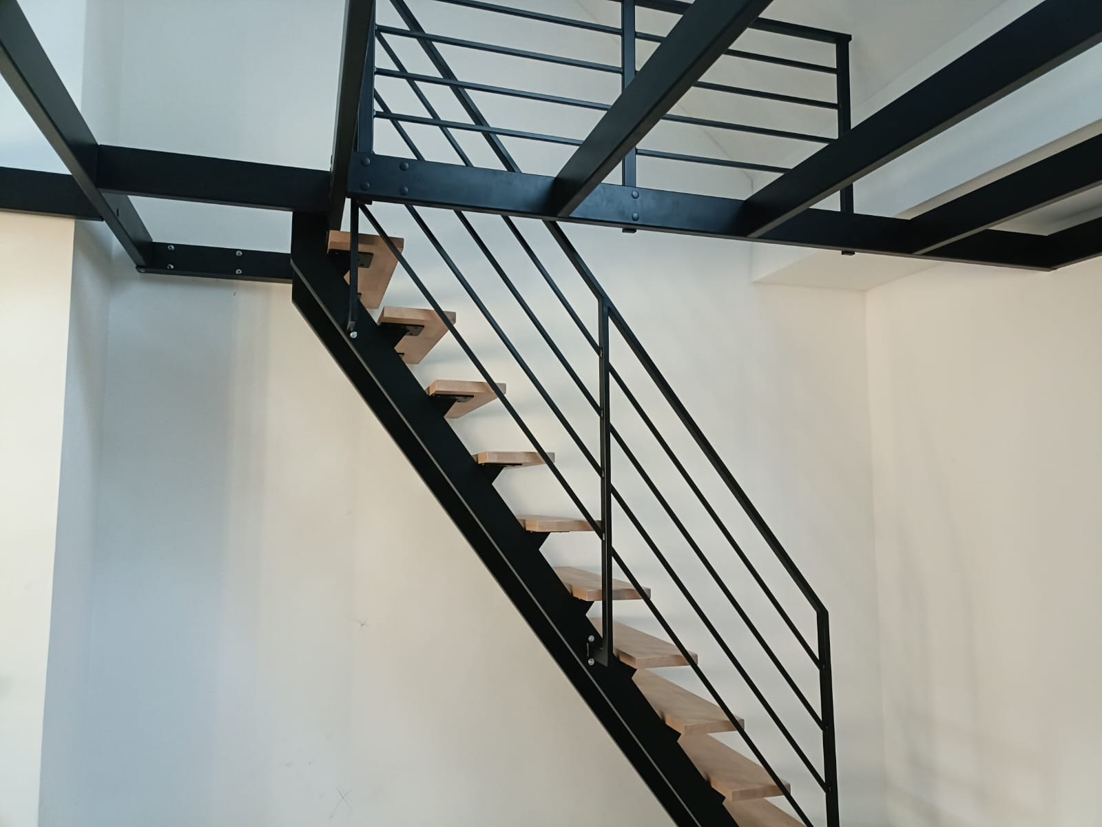 Nowoczesna antresola z czarną, metalową konstrukcją i drewnianymi stopniami schodów, widziana z dołu.