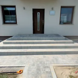 Schody wejściowe do domu, wyłożone szarą kostką brukową, prowadzące do drzwi wejściowych. W tle widoczny fragment elewacji domu z dwoma oknami. Obok leży łopata.