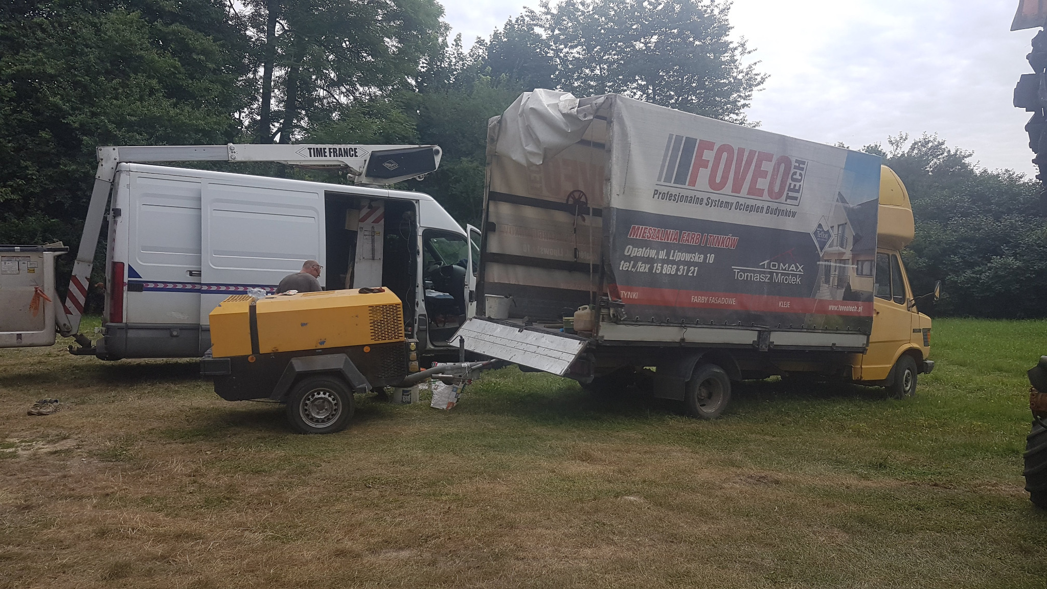 Dwa busy budowlane, biały z podnośnikiem koszowym Time France i żółty z reklamą Foveo-Tech, zaparkowane na trawiastym terenie, z widocznym kompresorem na przyczepce i pracownikiem wewnątrz białego...