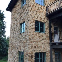 Kamieniarstwo Pawlik - Elewacja budynku wykończona kamieniem naturalnym o nieregularnym kształcie, z zielonymi ramami okiennymi i balkonem z czarną balustradą.