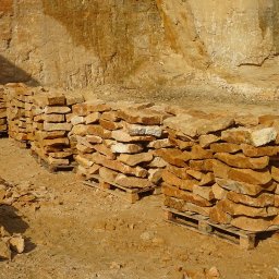 Kamieniarstwo Pawlik - Ułożone na paletach bloki kamienne w odcieniach beżu i brązu, widoczne na tle skalnej ściany w kamieniołomie, oświetlone mocnym, naturalnym światłem.