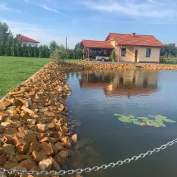 Brzeg oczka wodnego umocniony dużymi, nieregularnymi kamieniami w odcieniach brązu i beżu, z widocznym łańcuchem zabezpieczającym, w tle dom z czerwonym dachem i zielony trawnik pod błękitnym niebem.