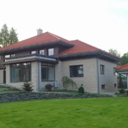 Nowoczesny dom jednorodzinny z elewacją z jasnego kamienia, czerwoną dachówką i zadbanym trawnikiem oddzielonym niskim murkiem z łupka.